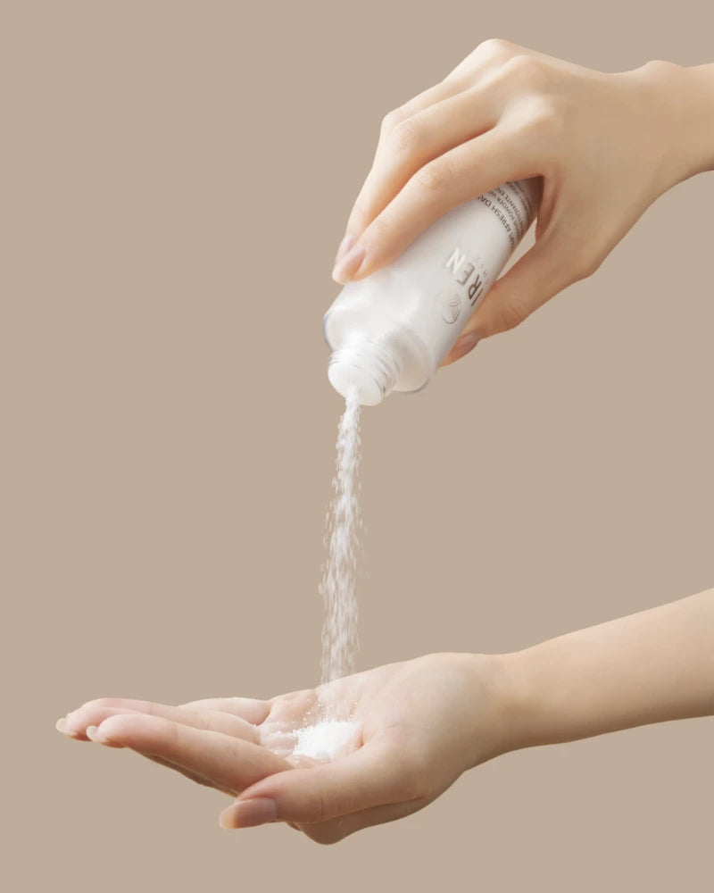 A person pours IREN Shizen’s START AFRESH DAILY Enzyme Powder Wash from a bottle onto their open palm, with a plain beige background.