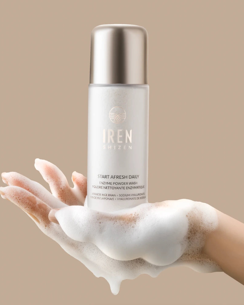 A foam-covered hand holds a bottle of IREN Shizen START AFRESH DAILY Enzyme Powder Wash—a gentle enzyme cleanser and daily exfoliant—against a beige background.