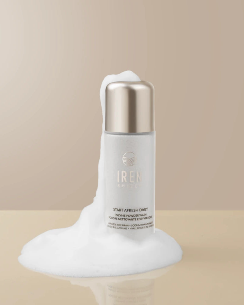 A white bottle of IREN Shizen START AFRESH DAILY Enzyme Powder Wash, a gentle exfoliant, is placed on a beige surface with white foam around it and a neutral background.