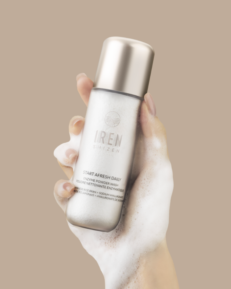 A hand holds a bottle from the FRESH AND CLEAR Cleansing Kit by Fast Bundle, covered in foamy lather, against a plain beige background—perfect for your skincare routine.