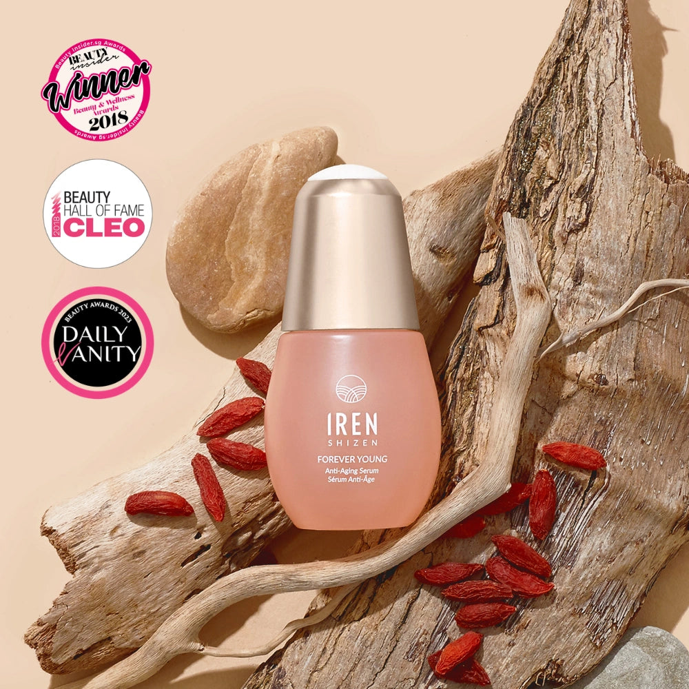 A bottle of IREN Shizen FOREVER YOUNG Anti-Aging Serum rests on driftwood with goji berries, framed by three award badges in the top left, emphasizing its anti-aging properties and collagen support.
