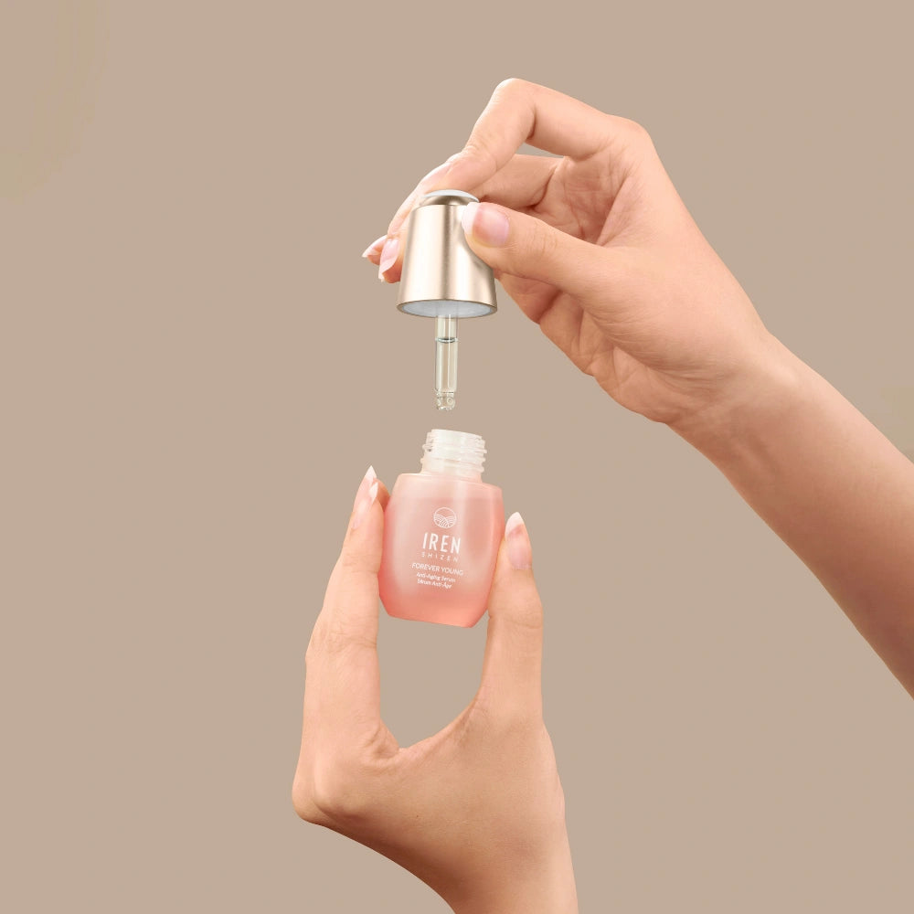 A person holds a dropper and a small glass bottle of pink FOREVER YOUNG Anti-Aging Serum by IREN Shizen, designed to support collagen production, against a plain beige background.