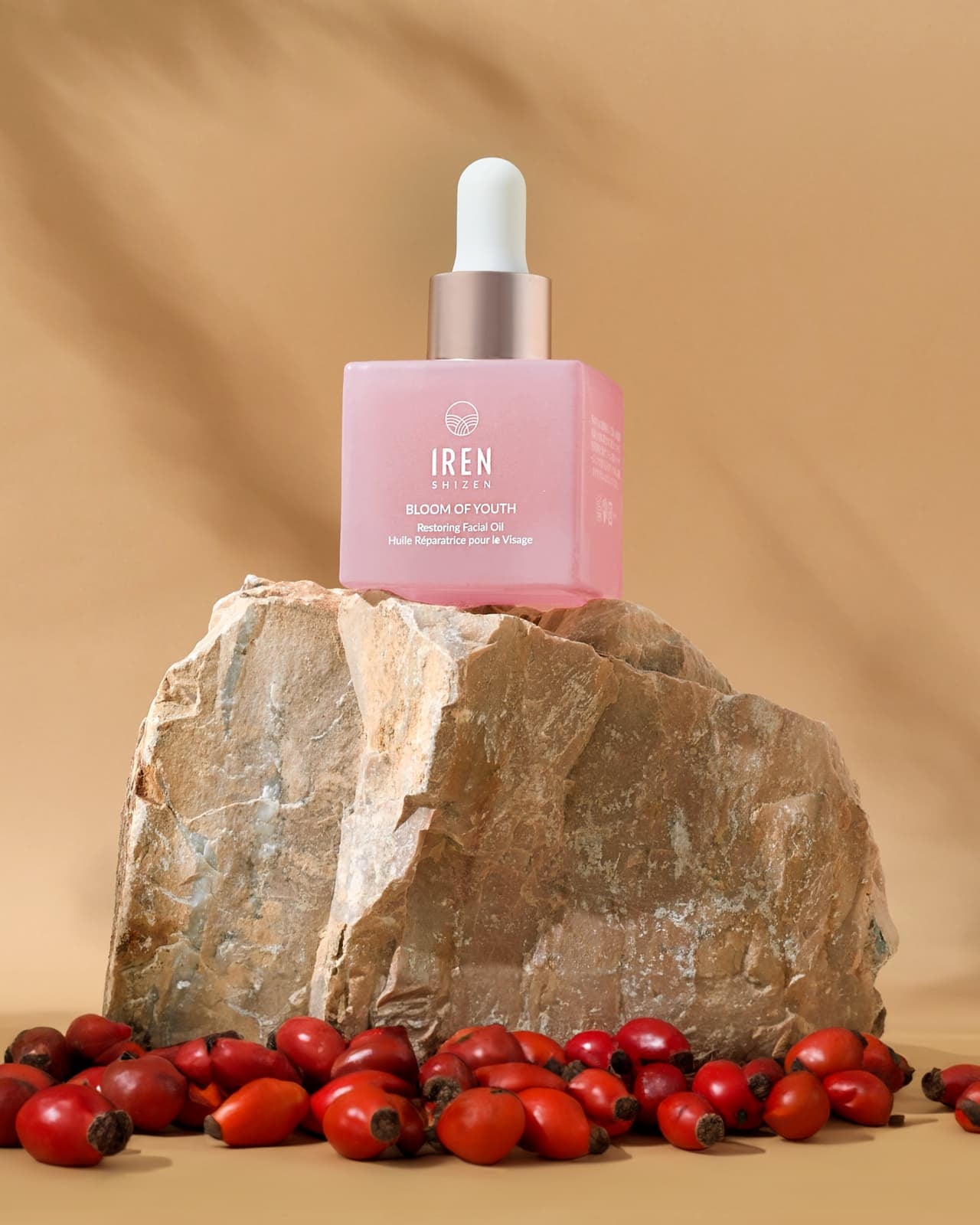 A pink dropper bottle labeled "IREN Shizen GLOW ON THE GO Travel Light Kit" sits on a rock, surrounded by scattered rose hips and a travel skincare kit against a beige background.