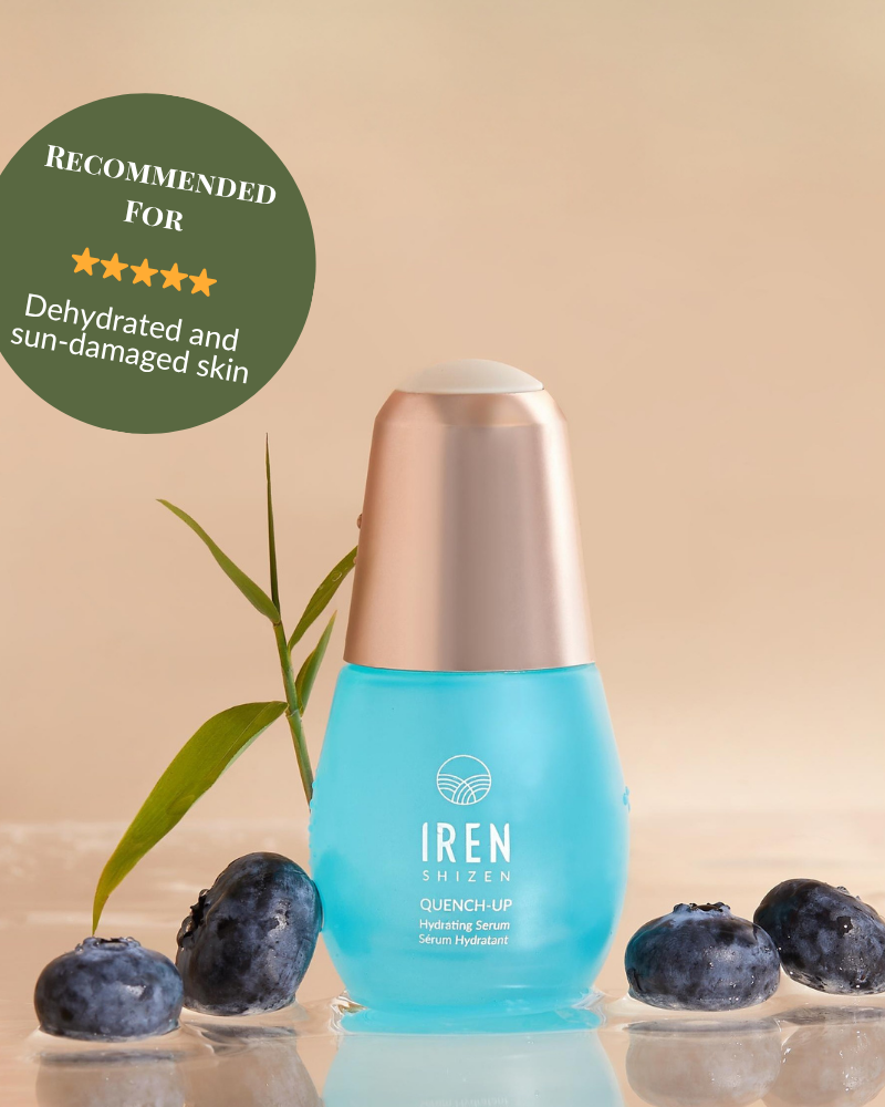A blue bottle of IREN Shizen QUENCH-UP Hydrating Serum with hyaluronic acid sits on a wet surface among blueberries and a green leaf, labeled for dehydrated and sun-damaged skin to help achieve plump skin.