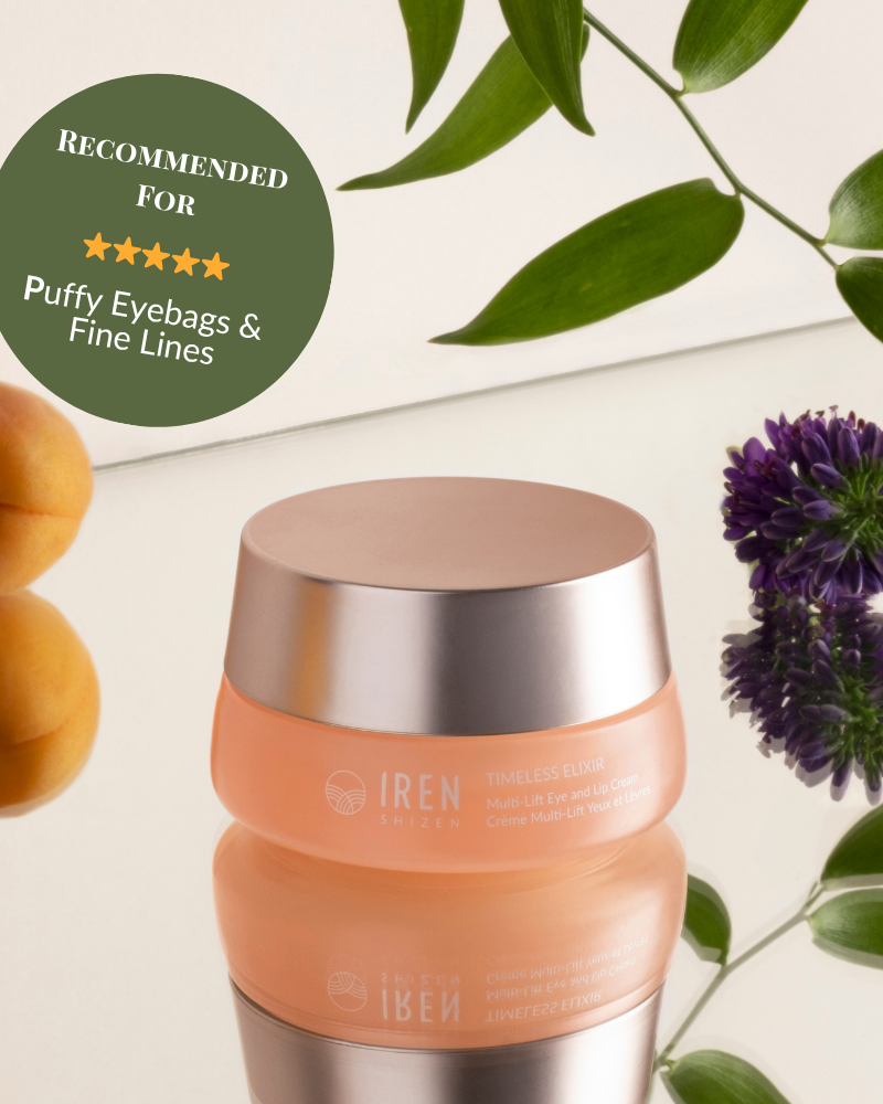 A peach IREN Shizen TIMELESS ELIXIR Multi-Lift Eye and Lip Cream jar sits on a reflective surface with leaves, purple flowers, and apricots. A green badge recommends it for treating puffy eyebags, fine lines, and dark circles. #show-lang-en