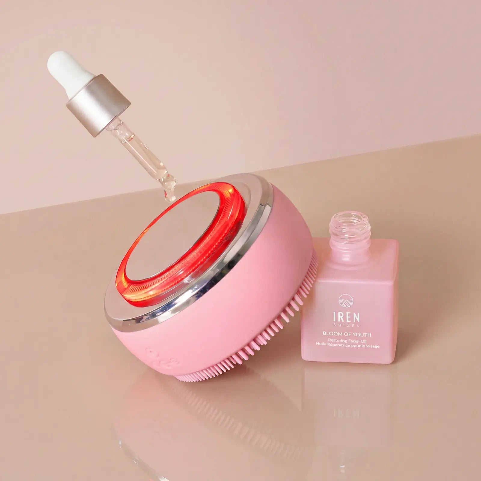 The IREN Shizen GLOW ON THE GO Travel Light Kit features a pink facial cleansing brush with LED light therapy beside a pink IRÉN Bloom of Youth serum bottle and dropper.