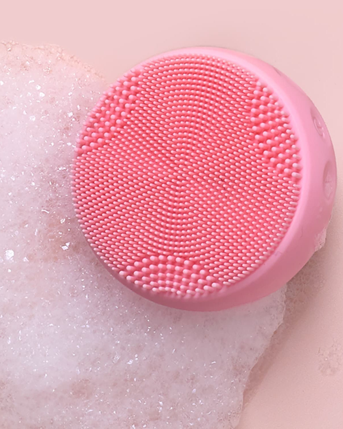 The GLOW ON THE GO Travel Light Kit by IREN Shizen features a round pink silicone cleansing device with small bristles, shown on foamy soap against a pale pink background—ideal for any travel skincare kit.
