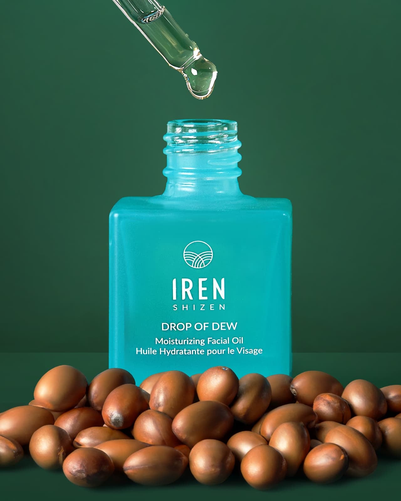 A dropper dispenses oil into a blue bottle labeled "IREN Shizen GLOW ON THE GO Travel Light Kit," surrounded by brown seeds on a green background—making it the perfect choice for any travel skincare routine.