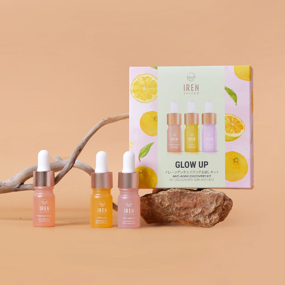 Three dropper bottles of IREN Shizen serums are shown with the GLOW UP Anti-Aging Discovery Kit box, accompanied by driftwood and citrus fruits on a beige background.