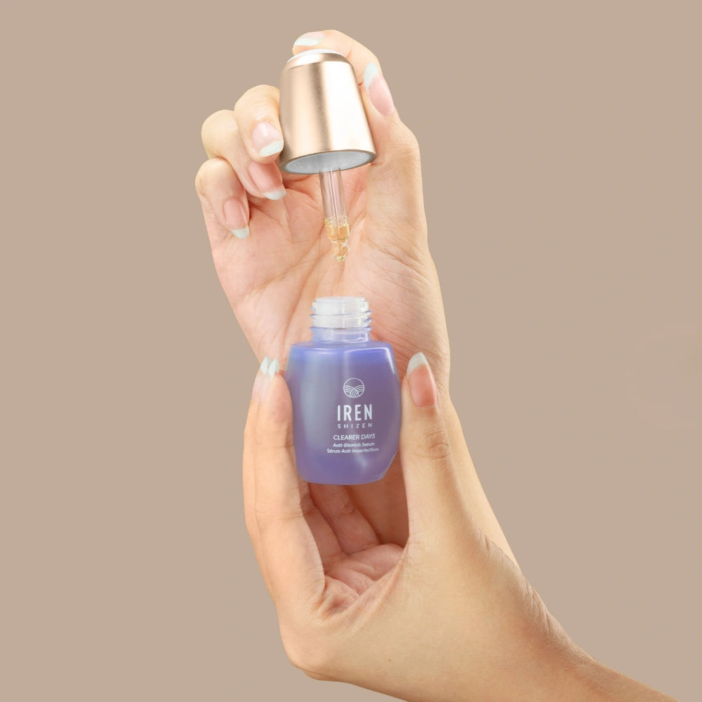 A hand holds a purple IREN Shizen CLEARER DAYS Anti-Blemish Serum bottle while another dispenses the serum with a dropper against a beige background, highlighting its acne treatment and sebum-balancing benefits. #all-groups