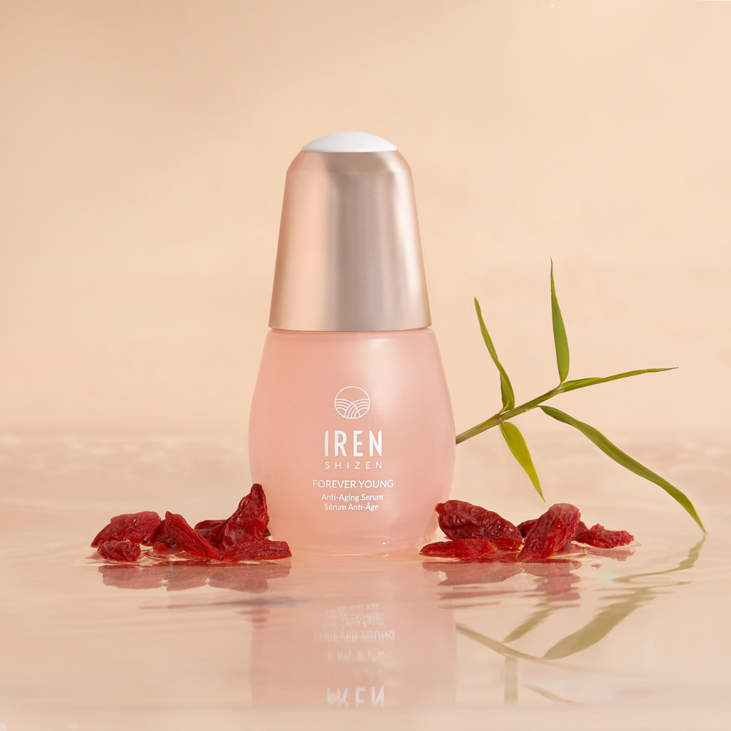 A bottle from the IREN Shizen TIMELESS ELIXIR Anti-Aging Eye Skincare Kit is displayed with scattered red berries and a sprig of green leaves on a reflective surface.