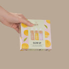 A hand holds a box of the IREN Shizen GLOW UP Anti-Aging Discovery Kit, featuring colorful citrus graphics and images of three serum bottles on the packaging.