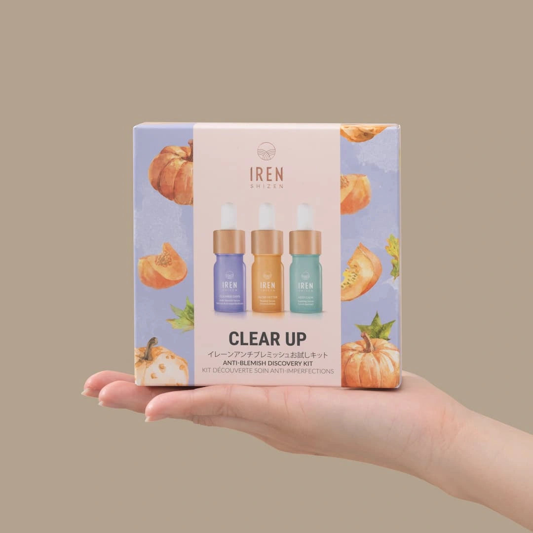 A hand holds a box of IREN Shizen CLEAR UP Anti-Blemish Discovery Kit, featuring three serums made with Japanese superfruits and decorated with pumpkins on a pastel background.