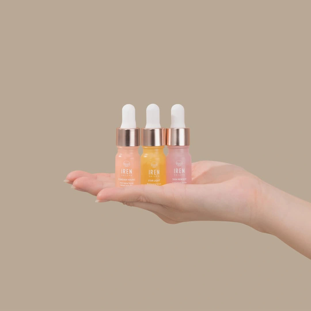 A hand holds three small dropper bottles from the IREN Shizen GLOW UP Anti-Aging Discovery Kit, featuring different colored serums against a plain beige background.