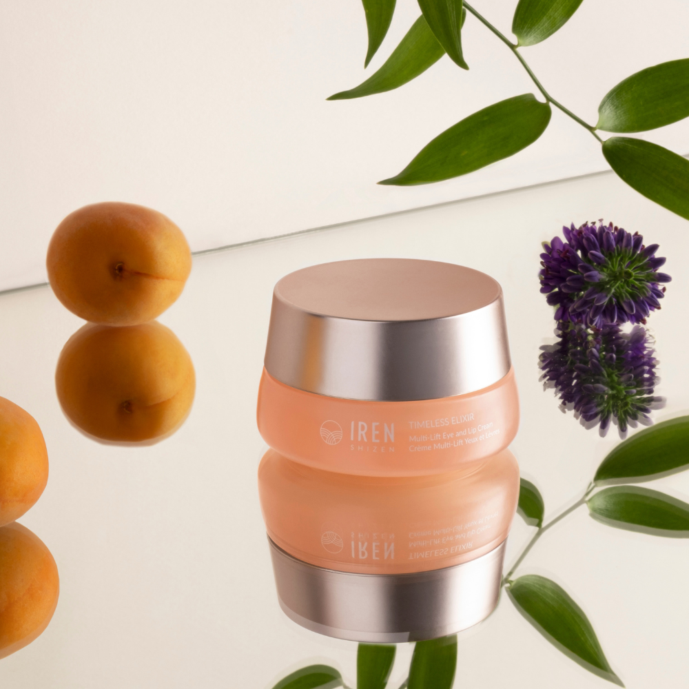 A jar of IREN Shizen TIMELESS ELIXIR Multi-Lift Eye and Lip Cream, celebrated for its anti-aging properties, is elegantly placed on a reflective surface surrounded by apricots, green leaves, and a purple flower.