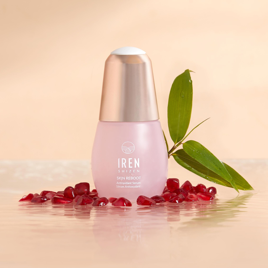 A bottle of TIMELESS ELIXIR Anti-Aging Eye Skincare Kit by IREN Shizen, a potent anti-aging eye treatment set, is placed upright, surrounded by scattered pomegranate seeds and a green leaf, set against a light peach background.