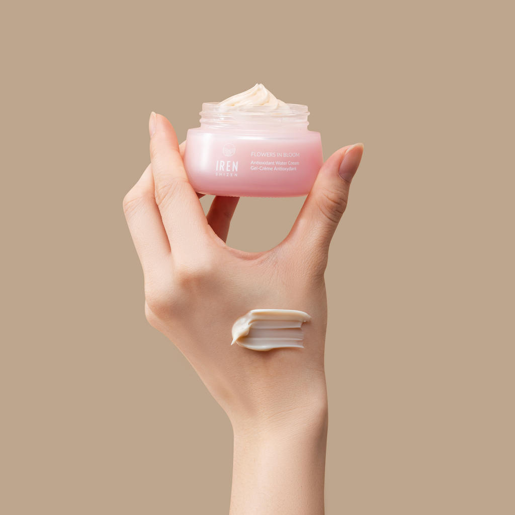 A hand holding an open jar of IREN Shizen's MOISTURE SANDWICH Skincare Kit with some product on the fingers against a neutral background.